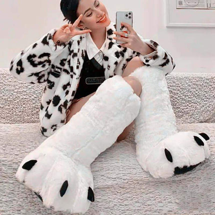 Cute Bear Claw Winter Boots. Person wearing large white paw-shaped slippers with a fluffy texture, sitting on a couch.