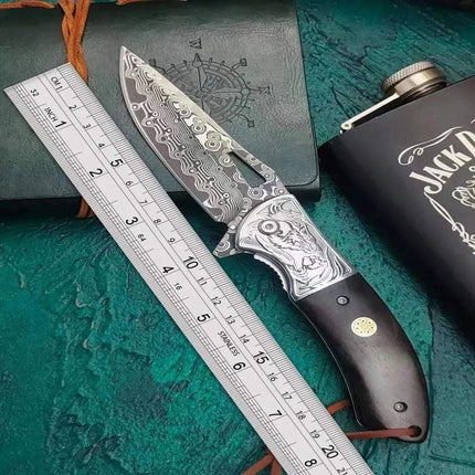 Damascus Steel Pocket Knife displayed with a ruler, leather-bound book, and flask on a textured background.