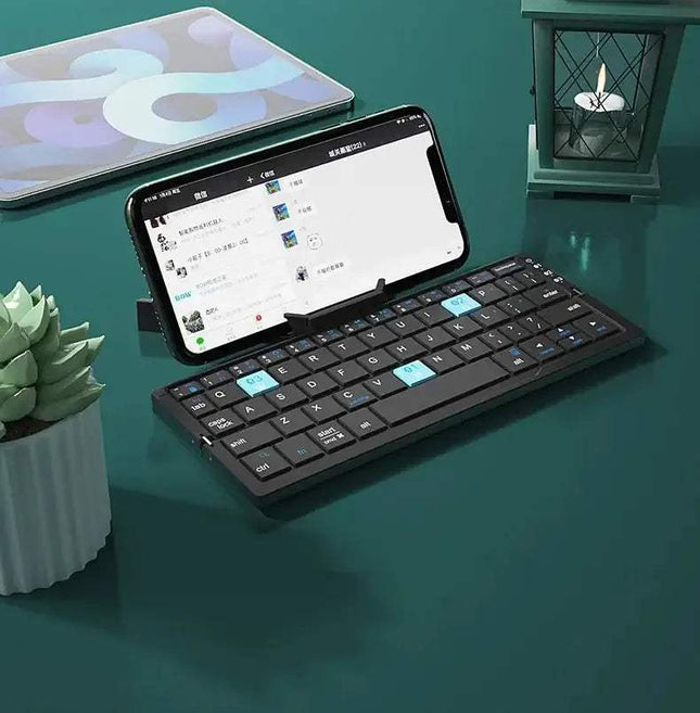 Mini Foldable Bluetooth Keyboard  attached to a smartphone on a teal surface with a plant and candle in the background.