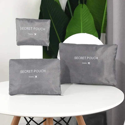 Three gray 6Pcs Travel Clothes Storage Bags 'SECRET POUCH' travel bags on a white surface with green leaves in the background.