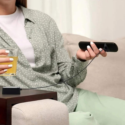Person sitting on a couch holding a glass of juice and a remote control with Bedside Remote Cable Lock Retractable Tether attached on a neutral background.