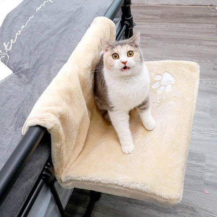 Cat sitting on a beige Cat Hanging Bed Plush Orthopedic Perch attached to a bed frame.